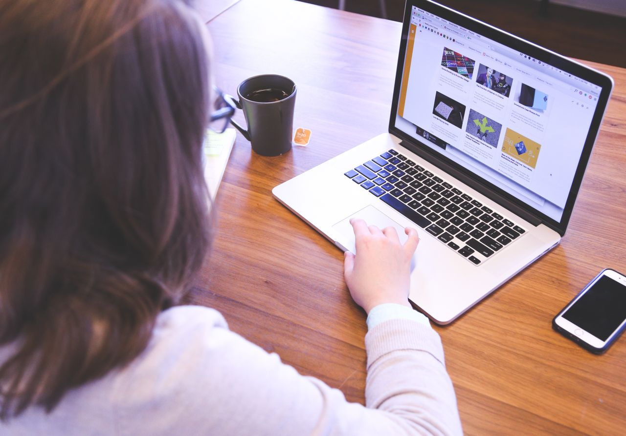 A person is using a MacBook to browse online while seated at a wooden desk, with a smartphone nearby.
